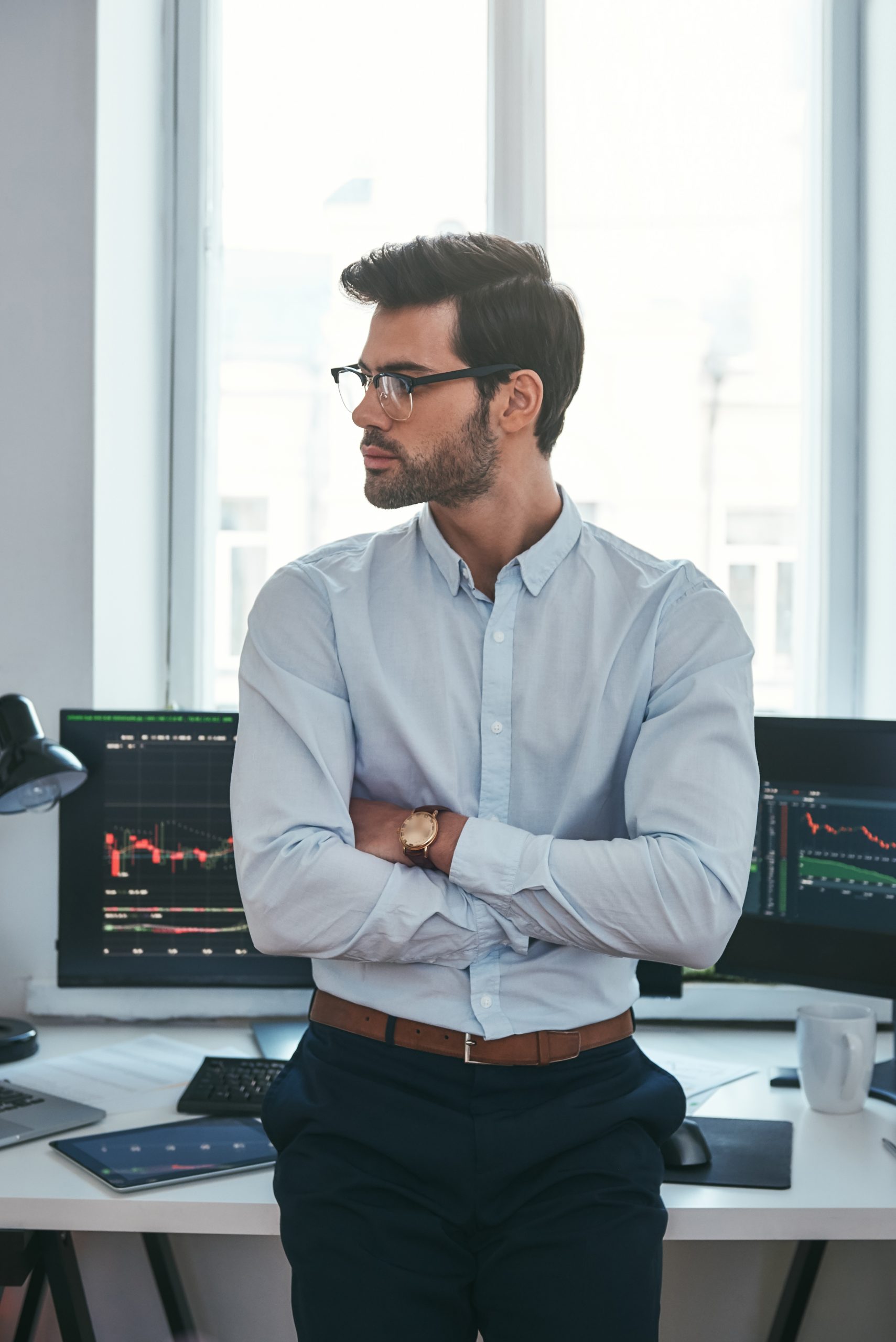 Young and smart. Vertical photo of confident businessman or trader in formal clothes and eyeglasses looking away and keeping arms crossed while standing at office.